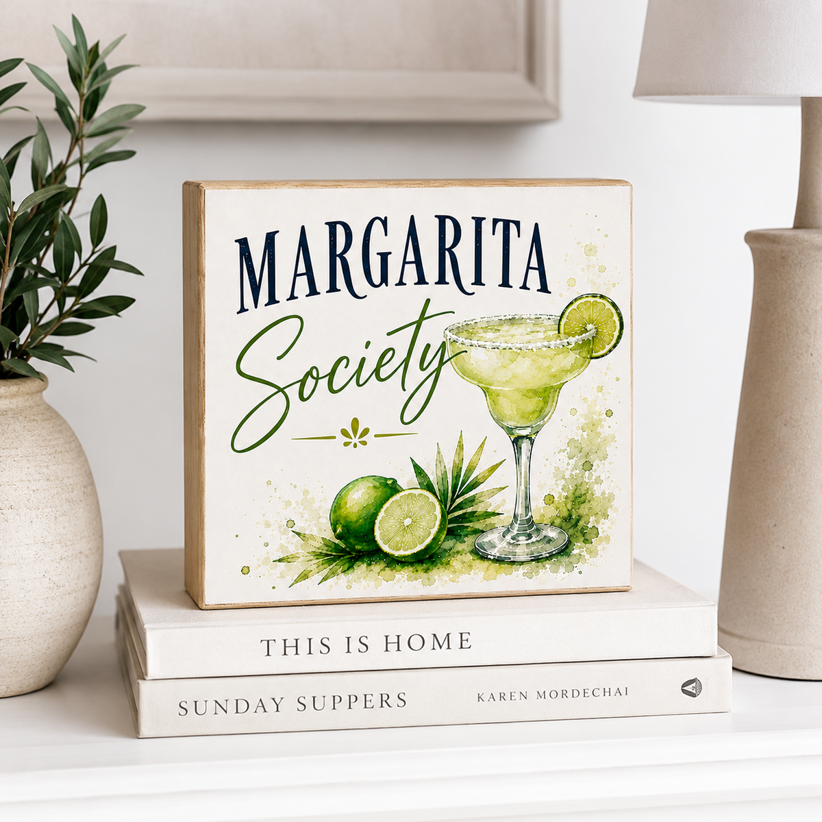 A square decorative sign reading “MARGARITA Society” with a watercolor-style margarita glass, lime, and greenery illustration, displayed on stacked books in a styled interior setting.