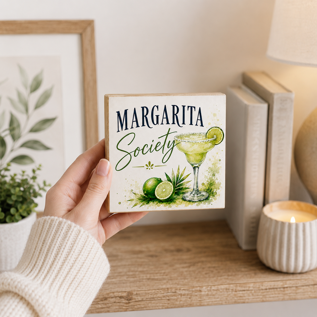 A hand wearing a cream knit sweater holds a square wooden coaster featuring a watercolor-style illustration of a margarita glass, lime slices, and the words “MARGARITA Society.” The background shows a warm, styled interior shelf with books, a candle, and framed botanical art.