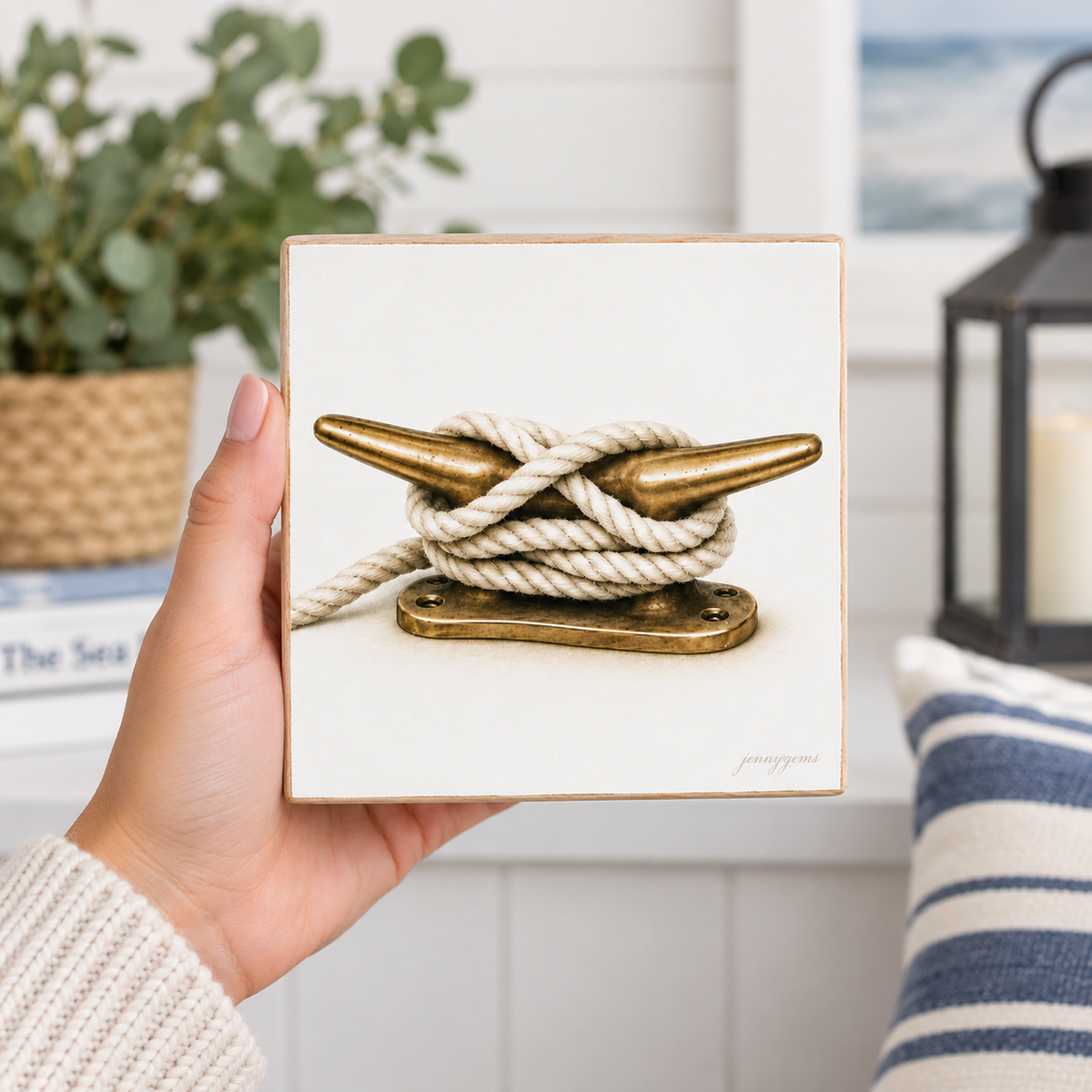 A hand holds a square white canvas print featuring a nautical art piece of a brass anchor tied with white rope. The background shows a softly blurred nautical-themed interior with a plant, a striped pillow, and a lantern.