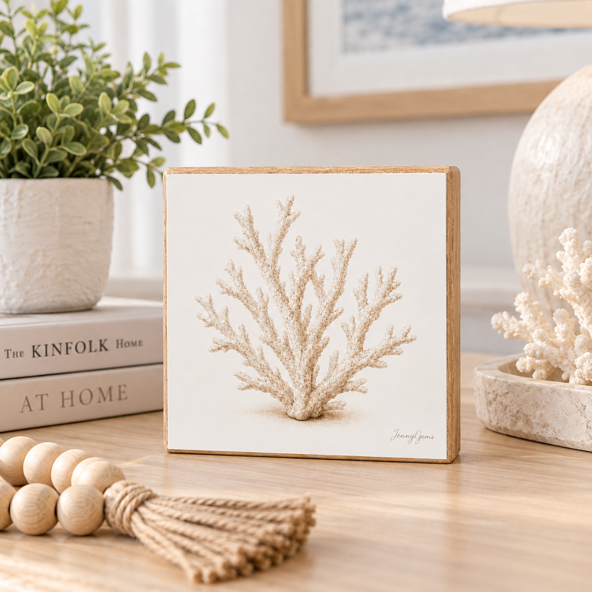 A square decorative block print featuring a beige coral illustration on a white background, styled on a light wood tabletop with a plant, books, and rope decor nearby.