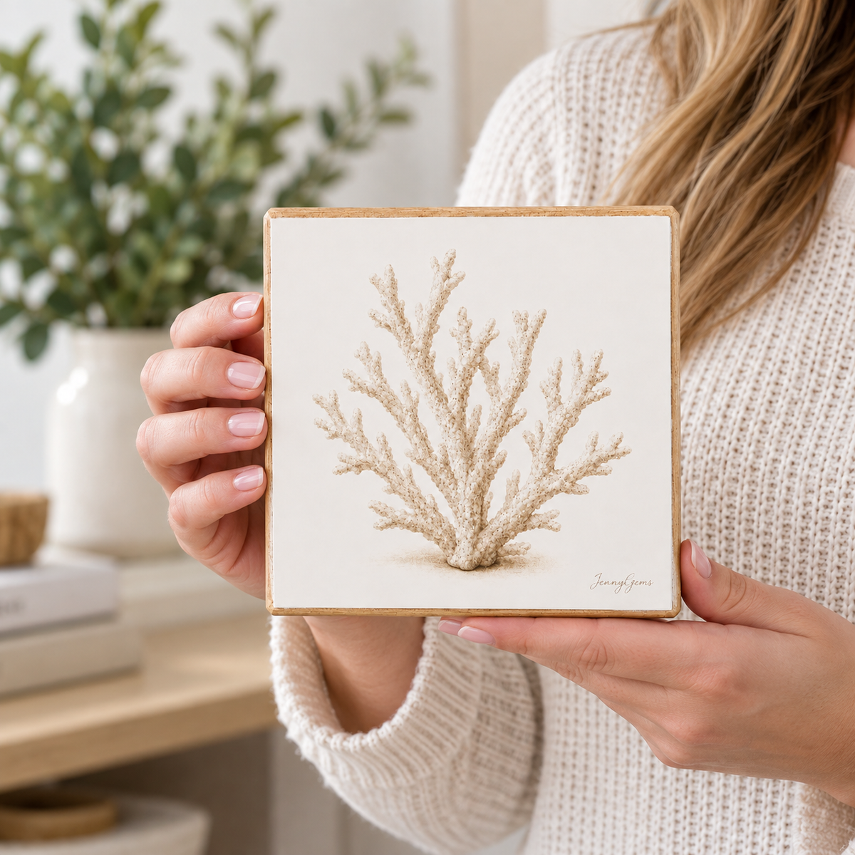 A person in a cream knit sweater holds a square ceramic tile featuring a hand-painted beige coral design, set against a softly blurred home background with a plant.