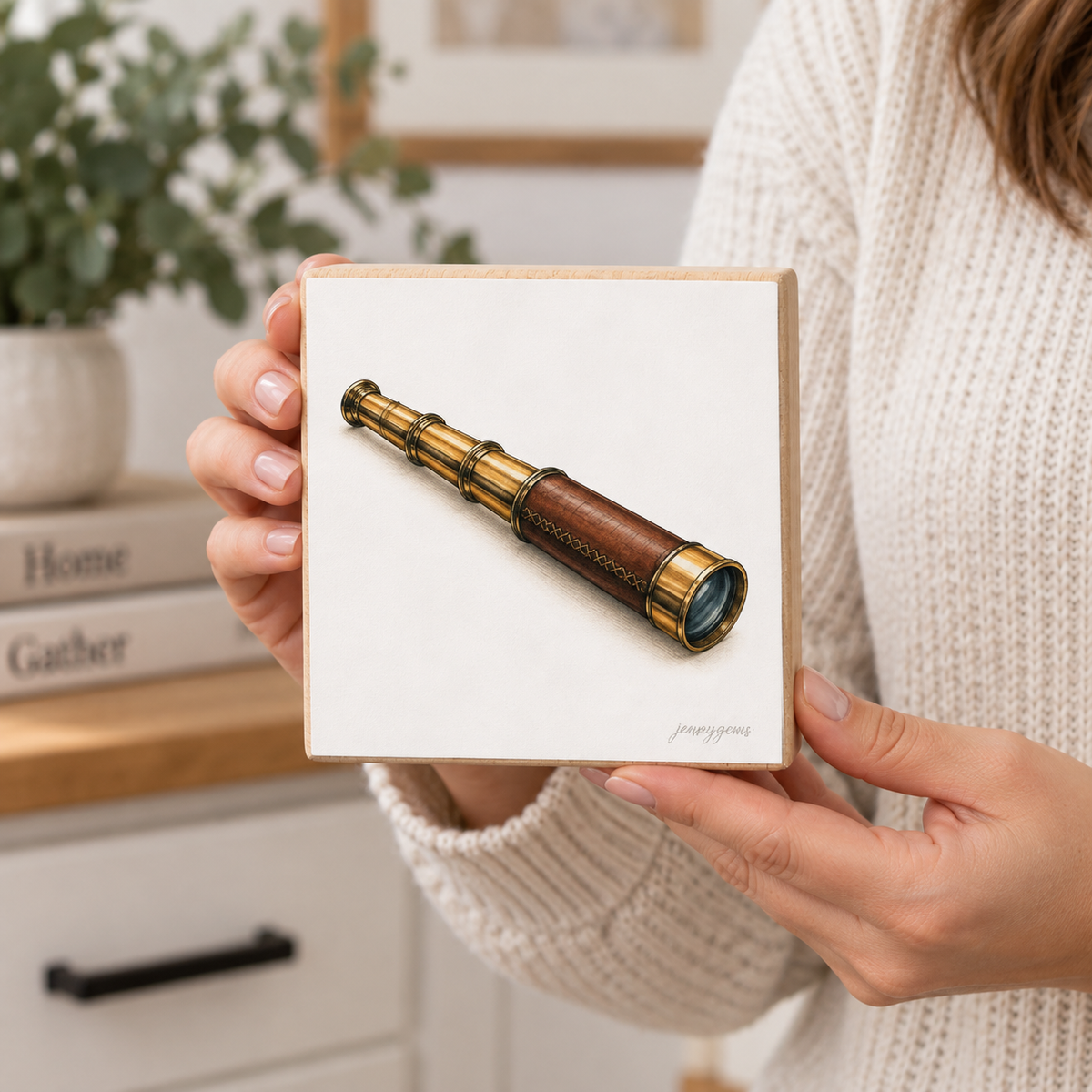 A person in a cream knit sweater holds a square white art print featuring a detailed illustration of a brass and brown telescope. The background shows a softly blurred home interior with a plant and books.