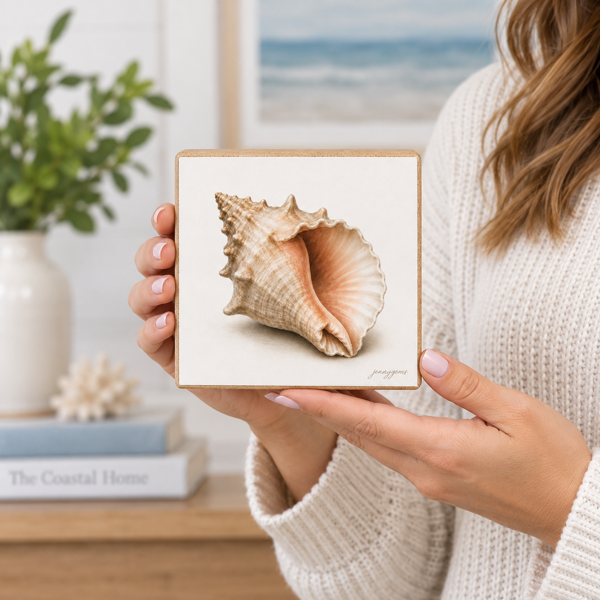 A person in a cream knit sweater holds a square art print featuring a detailed seashell illustration. The print has a light wood border and is shown in a cozy, styled indoor setting with a blurred background of a plant and framed ocean view.