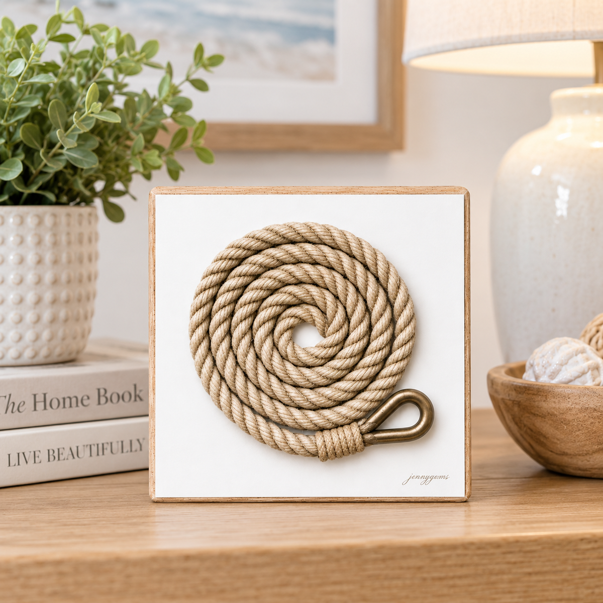 A coiled beige rope with a brass ring displayed on a white square card inside a light wood frame, set on a wooden table in a styled home decor setting with a plant, lamp, and books.