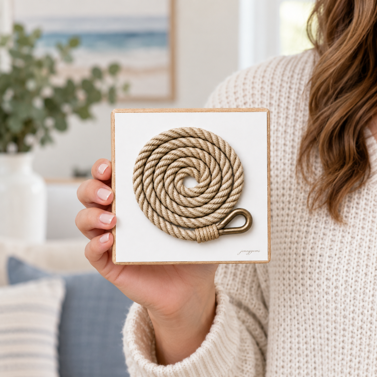 A person in a cream knit sweater holds a square white presentation box featuring a coiled beige rope with a brass-toned loop on a white background.