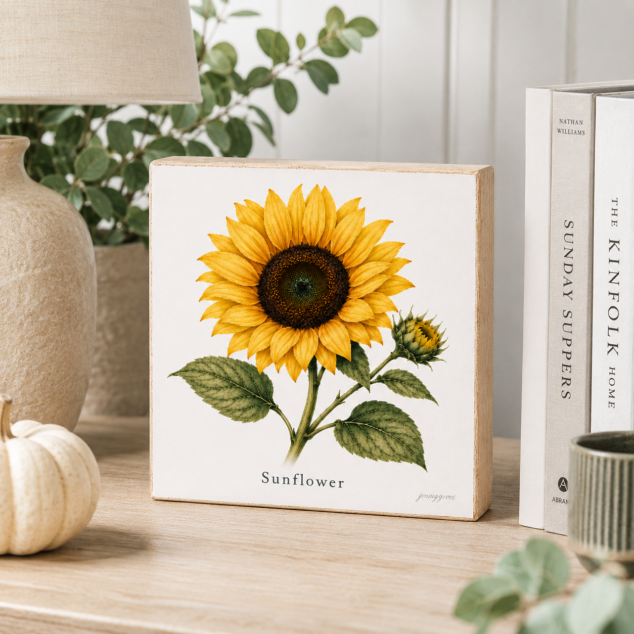 A square decorative block sign featuring a detailed sunflower illustration, styled on a wooden tabletop next to a ceramic lamp base, a small white pumpkin, books, and green foliage.