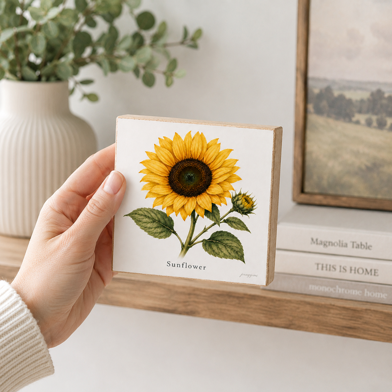 A hand holds a square wooden block print featuring a detailed sunflower illustration, set against a cozy home interior background with a vase of greenery and framed art.