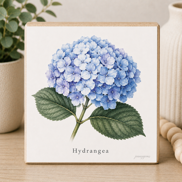 Framed artwork of a blue hydrangea flower on a wooden surface with a plant and vase in the background.