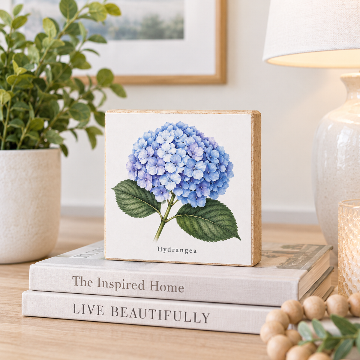 A square decorative block sign featuring a watercolor-style blue hydrangea with green leaves and the word 'Hydrangea' printed below, displayed on stacked books on a tabletop in a styled home setting.