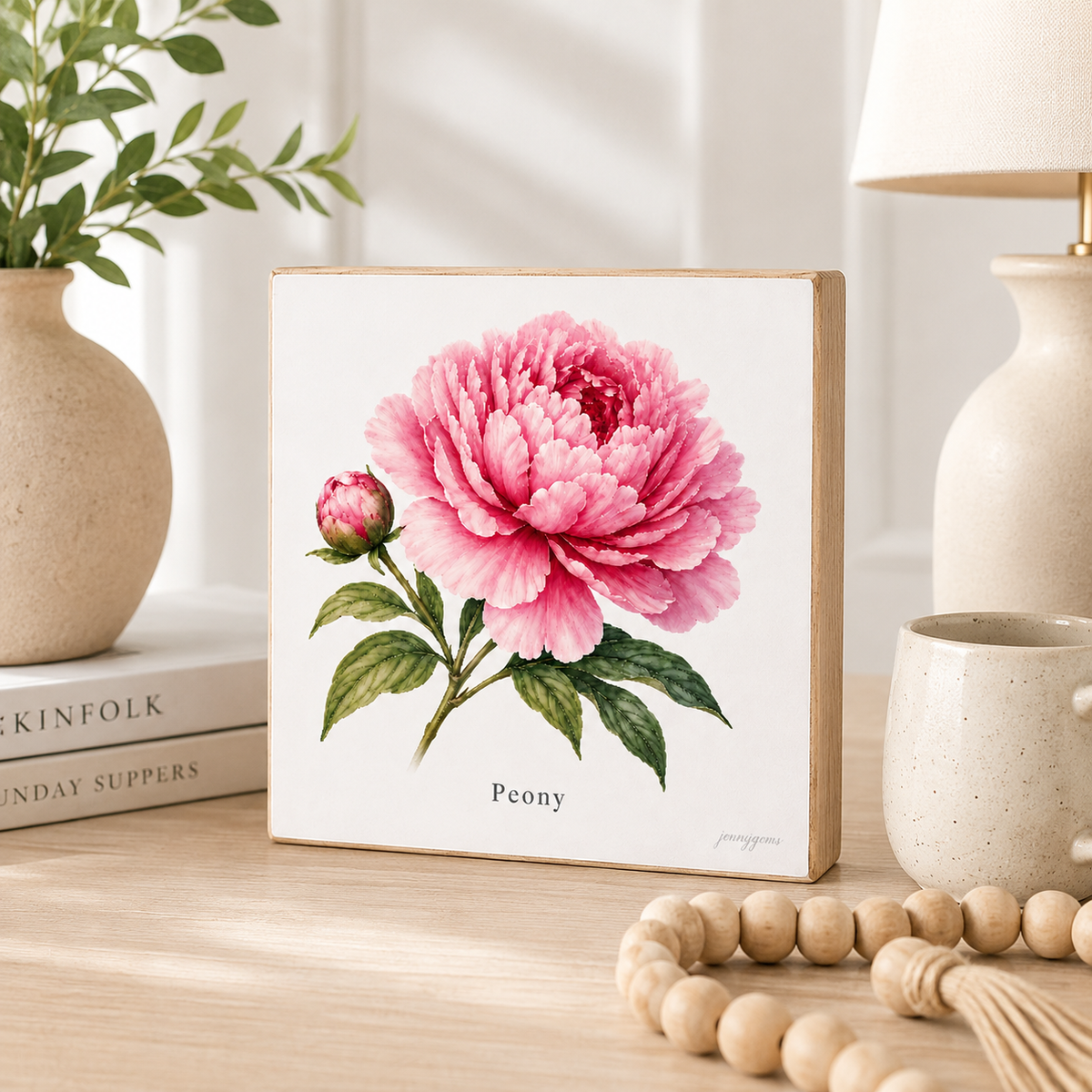 A square decorative block or plaque featuring a painted pink peony flower with green leaves on a white background, standing on a wooden table in a styled interior setting with books, a vase, lamp, and beads nearby.