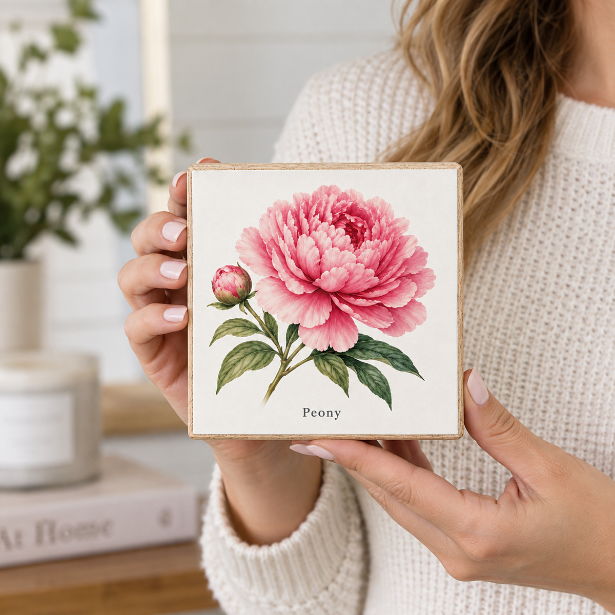 A woman in a cream knit sweater holds a square wooden box featuring a watercolor-style pink peony flower with green leaves and the word 'Peony' printed below. The background is softly blurred, showing a home interior with a plant and candle.