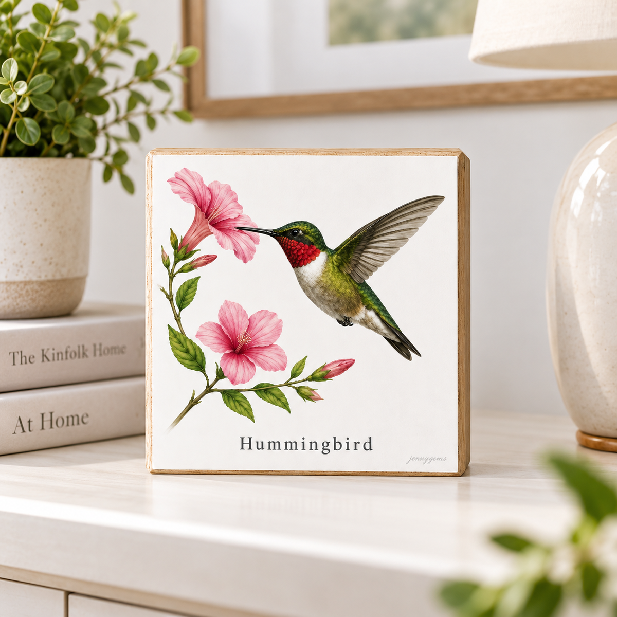A square wooden sign featuring a painted hummingbird with pink flowers and the text 'Hummingbird,' displayed on a tabletop in a styled home setting with books, a plant, and a lamp.