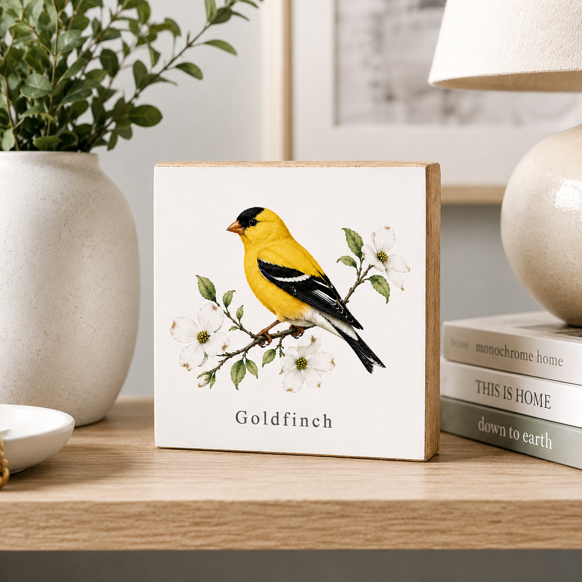 A square wooden plaque featuring a vintage-style illustration of a goldfinch bird perched on a flowering branch, with the word 'Goldfinch' printed below. The plaque is styled on a wooden shelf next to a ceramic vase with greenery, a stack of books, and a lamp.