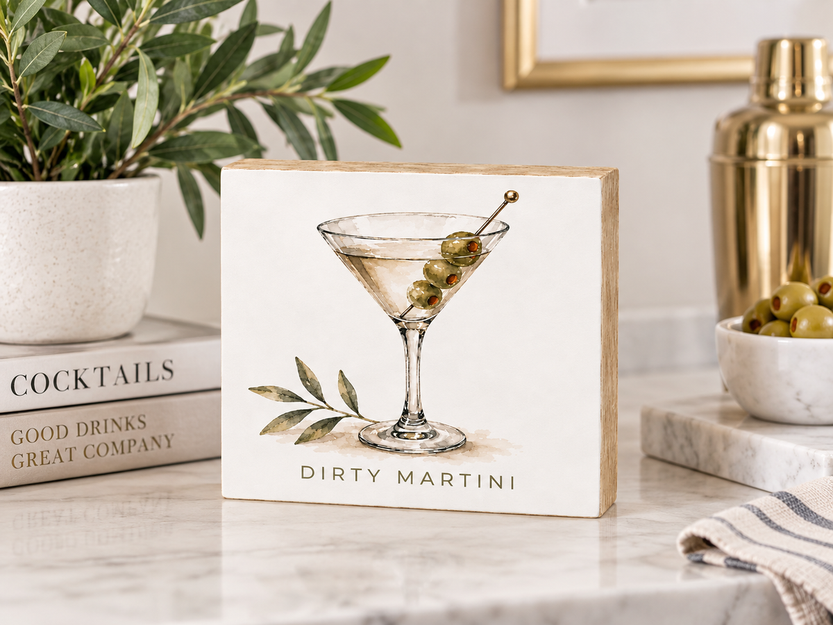 A square decorative sign with a light wood border and a white center featuring a watercolor-style martini glass with olives and the text 'DIRTY MARTINI,' styled on a marble countertop with books, a plant, and a cocktail shaker in the background.
