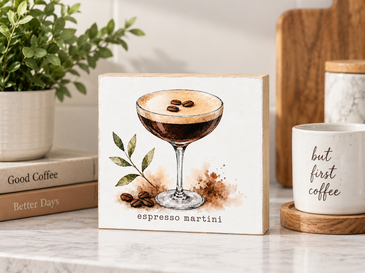 A square wooden sign featuring a watercolor-style espresso martini illustration, displayed on a marble countertop in a styled kitchen setting with a plant, books, and a mug nearby.