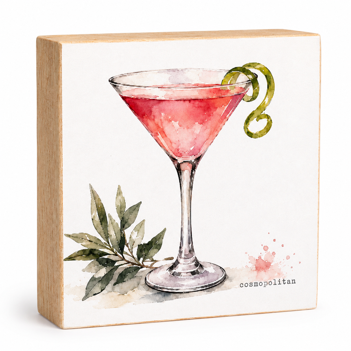 A square wooden block sign featuring a watercolor illustration of a pink martini cocktail in a coupe glass, garnished with a green twist and accompanied by green leaves. The text 'cosmopolitan' is printed in black near the bottom right.