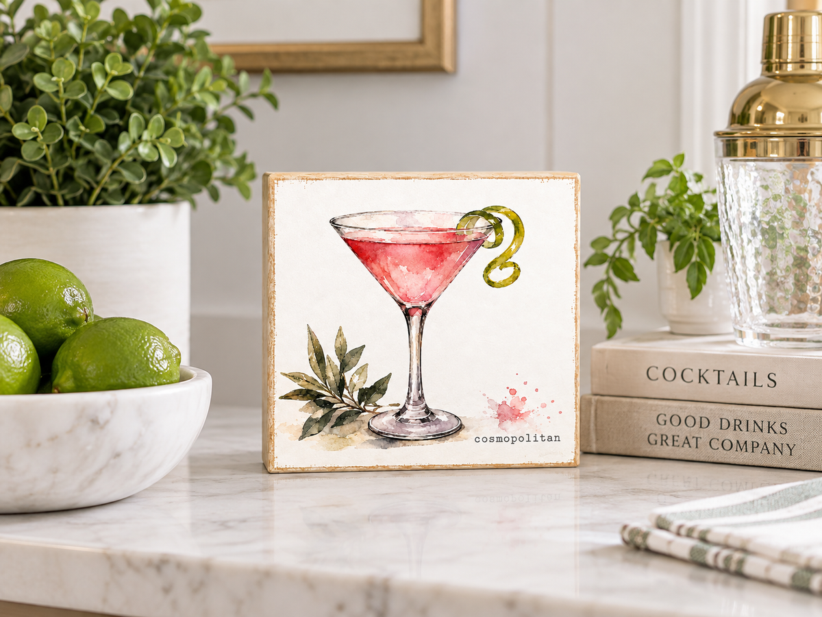 A square decorative tile featuring a watercolor-style pink martini cocktail with olives, displayed on a marble countertop in a styled kitchen setting with limes, plants, and books.