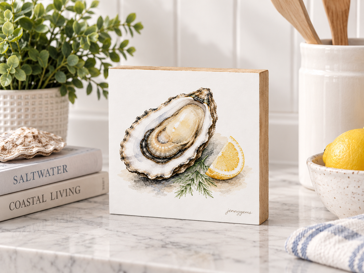 A square wooden sign featuring a watercolor illustration of an open oyster with a lemon wedge and dill, displayed on a marble countertop in a kitchen setting.