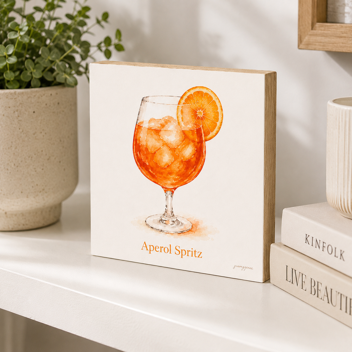 A square canvas art print featuring a watercolor illustration of an Aperol Spritz cocktail with an orange slice garnish, displayed on a white shelf next to a potted plant and books.