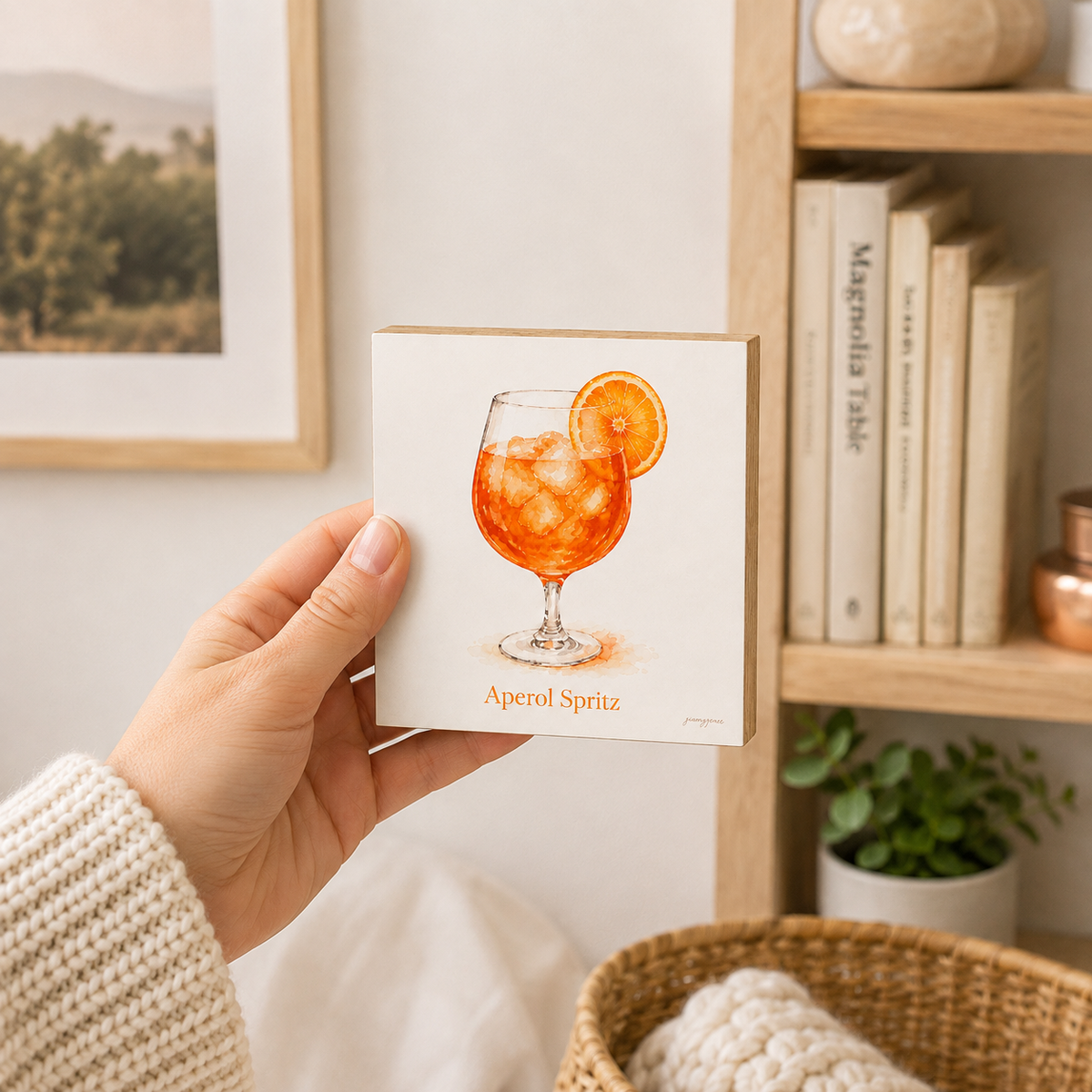 A hand holds a square art print featuring an orange-colored Aperol Spritz cocktail illustration with an orange slice garnish. The print is shown in a cozy indoor setting with a wooden bookshelf in the background.