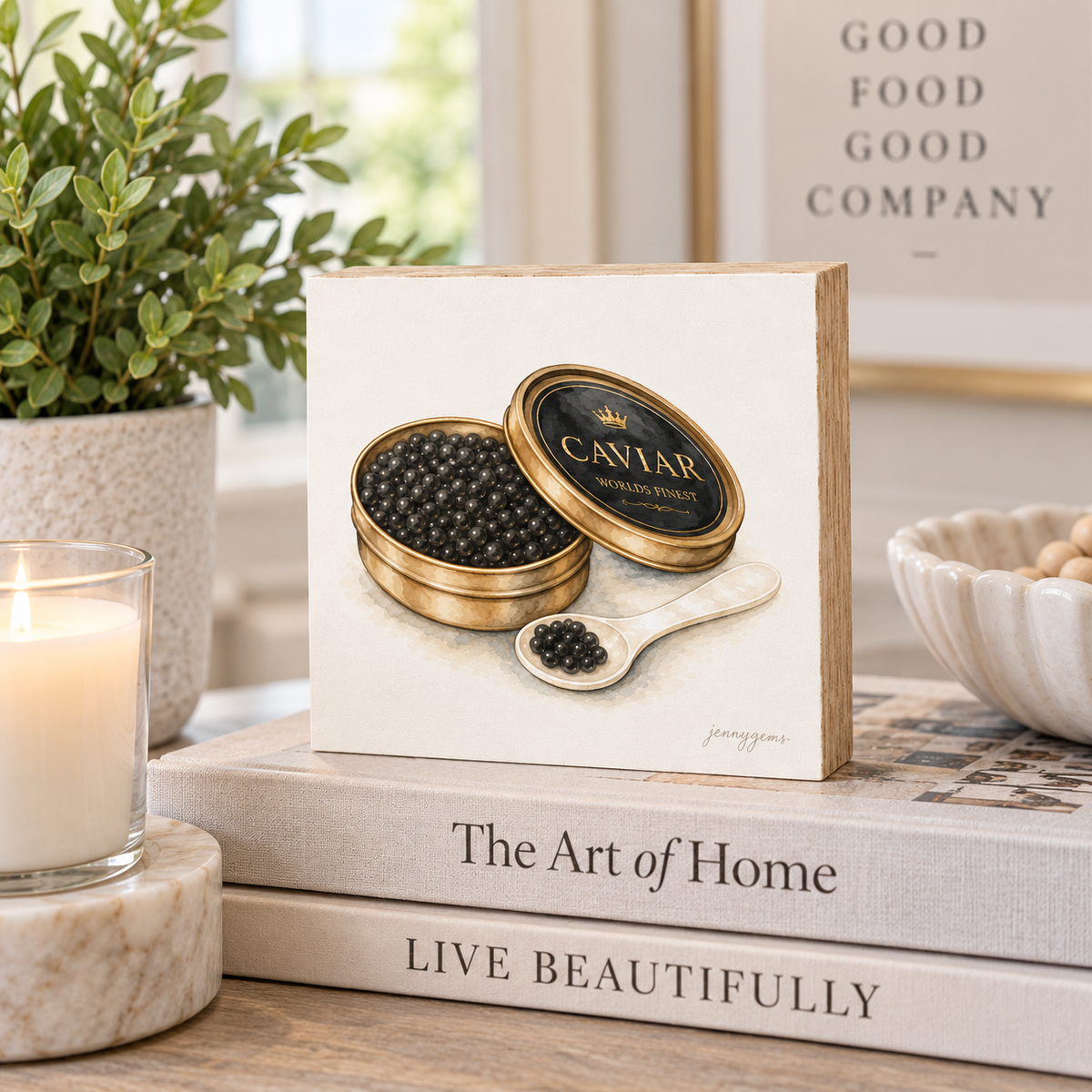 A square decorative print or box featuring an illustration of a gold caviar tin and spoon on an off-white background, styled on top of books in a warm home setting with a candle, plant, and bowl.