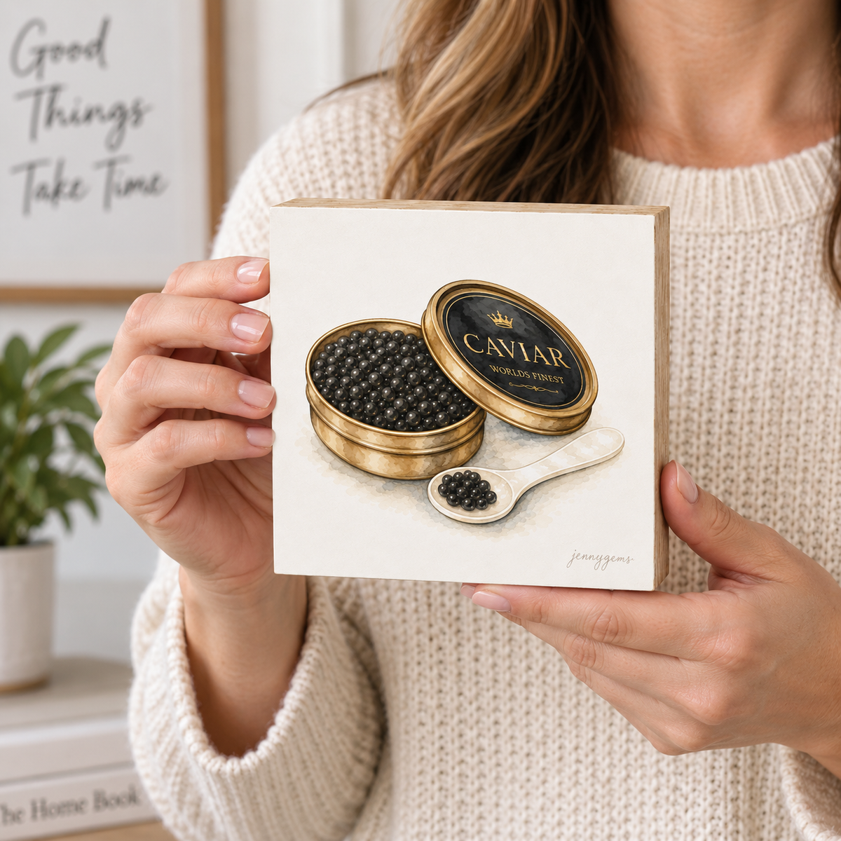 A woman in a cream knit sweater holds a square cream-colored art print or card featuring a painted illustration of an open gold caviar tin, a spoonful of black caviar, and the text 'CAVIAR WORLD'S FAVORITE.' The scene is indoors with a softly blurred background including framed wall art, a plant, and a book.