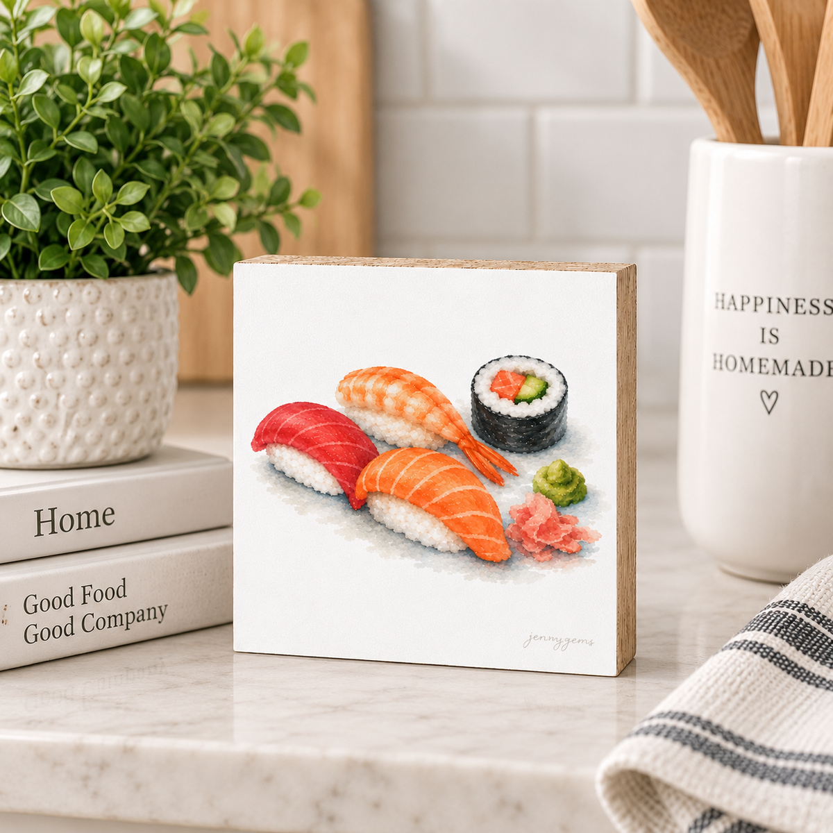 A square wooden block sign featuring a watercolor illustration of sushi, styled on a marble countertop with a potted plant, books, and kitchen utensils in the background.