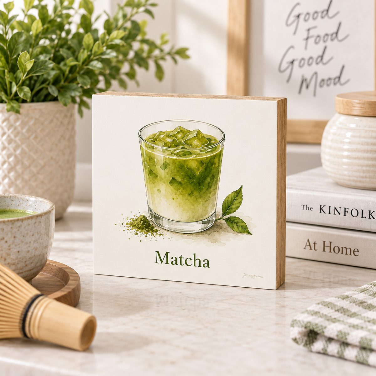 A square decorative wall art print or canvas featuring an illustration of a green matcha drink in a glass, with the word "Matcha" below. The artwork is displayed on a light marble countertop in a styled kitchen setting with a plant, books, a bowl, and a whisk nearby.