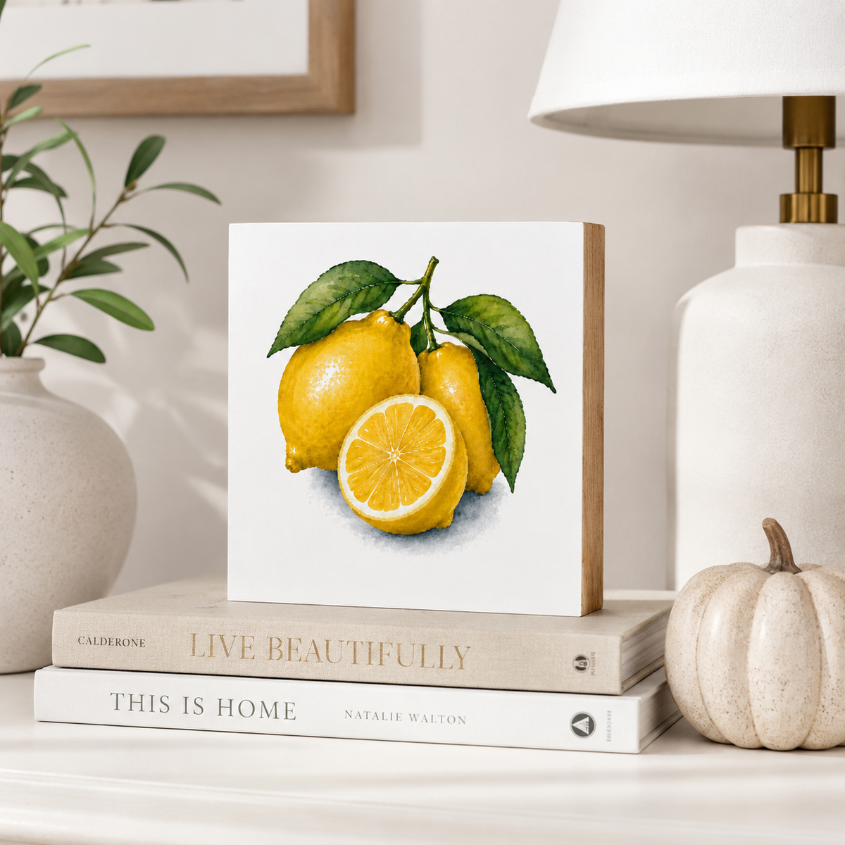 A square white art print or canvas featuring a watercolor-style illustration of three yellow lemons with green leaves, displayed on top of stacked books on a tabletop. A lamp, plant, and small pumpkin decor are visible in the styled room setting.