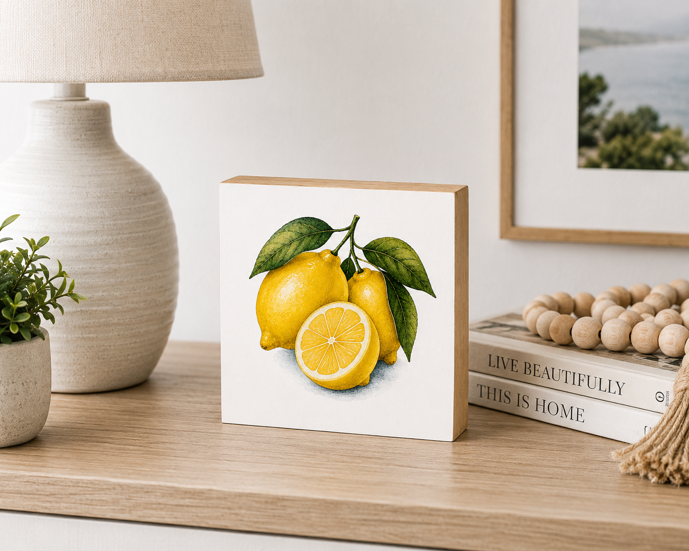 A square white art block featuring a watercolor-style illustration of three yellow lemons with green leaves, displayed on a wooden shelf next to a lamp, books, and decorative beads.