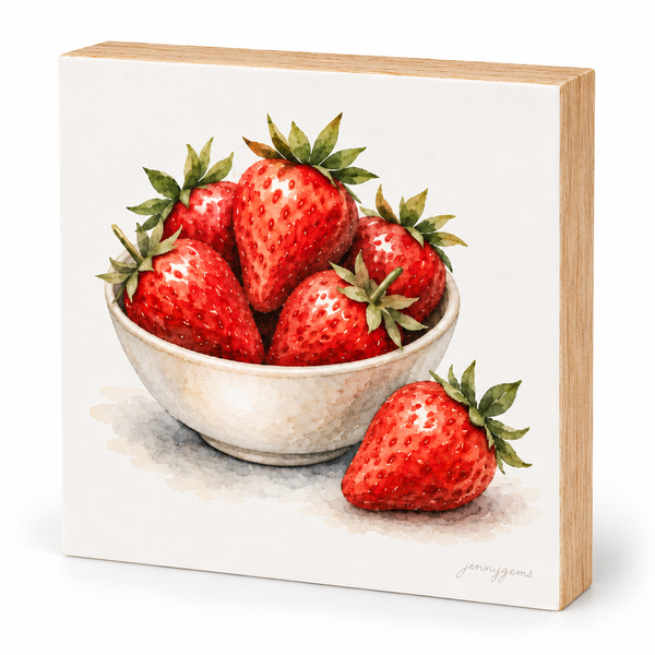 A square wooden block plaque featuring a watercolor-style painting of red strawberries in a white bowl, with one strawberry placed outside the bowl on the front. The wood side edge is visible, and a small artist signature appears in the lower right corner.