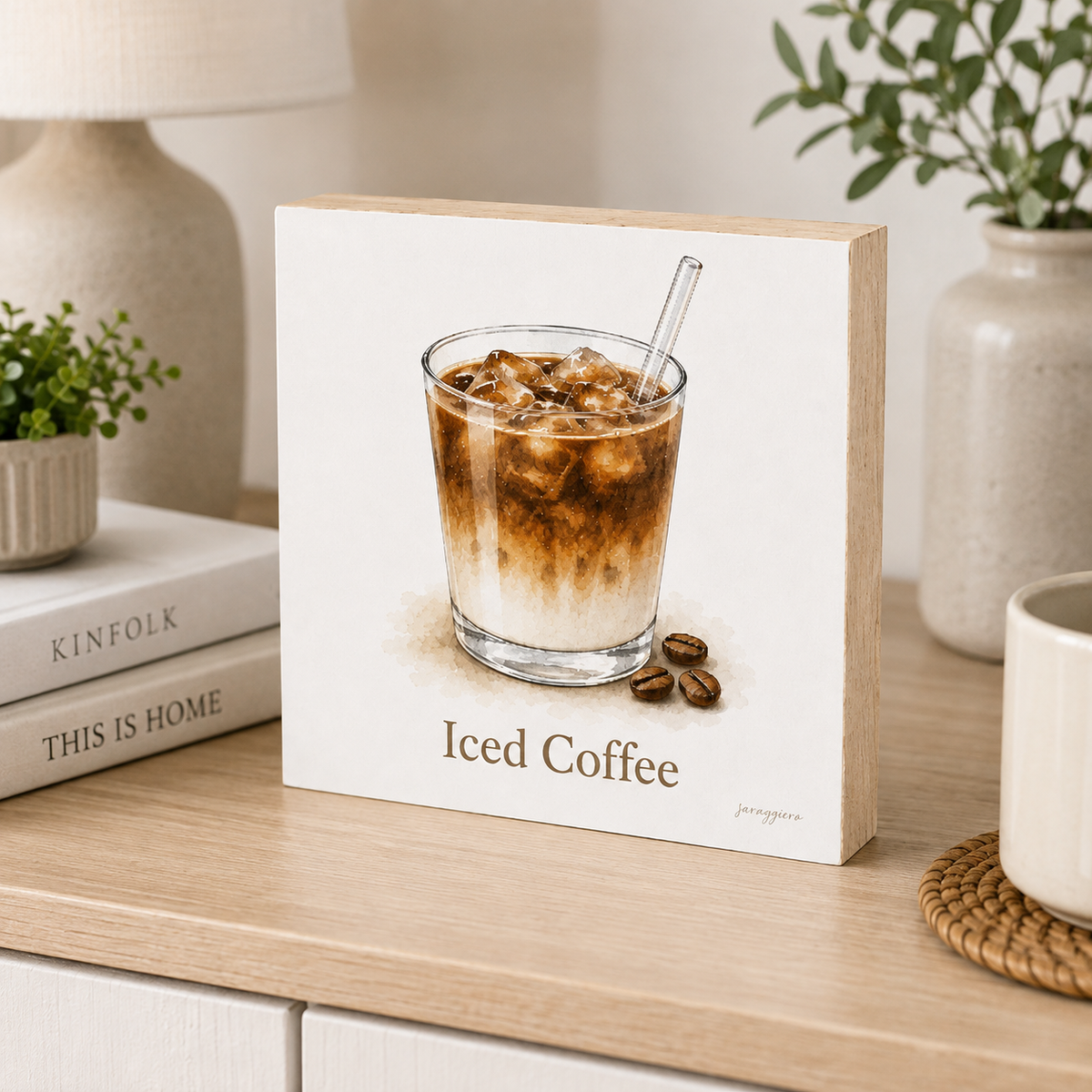A square decorative sign with a light wood border and a white face showing an illustration of an iced coffee drink in a glass, with the words 'Iced Coffee' and a small signature. The sign is styled on a light wood tabletop with books, a lamp, and a plant in the background.