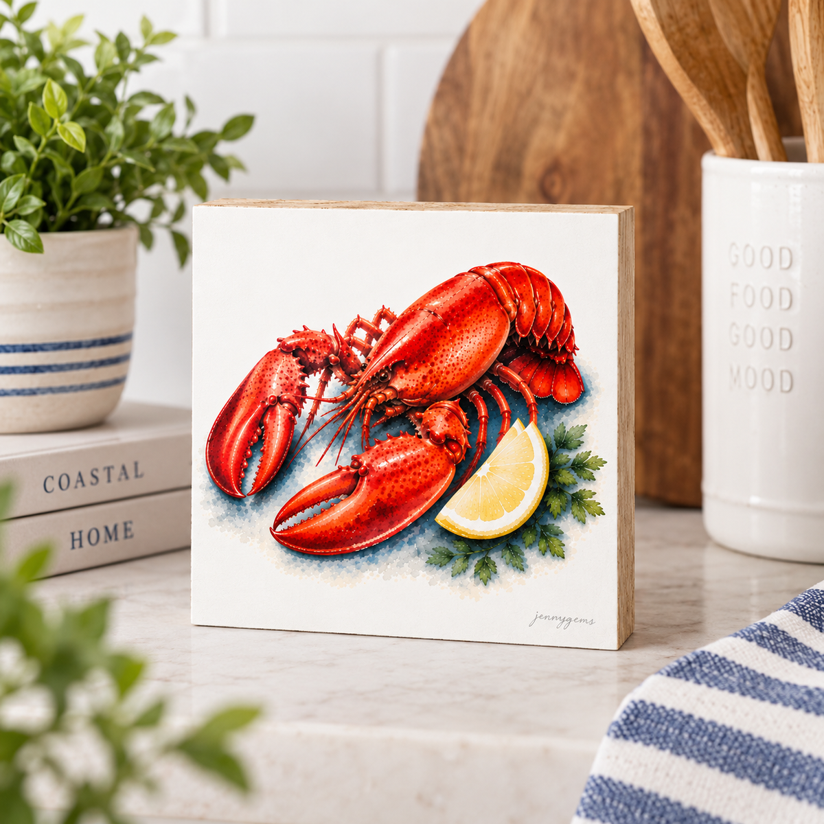 A square decorative wall sign featuring a painted lobster and lemon slice, displayed on a kitchen countertop with plants, books, and utensils in the background.