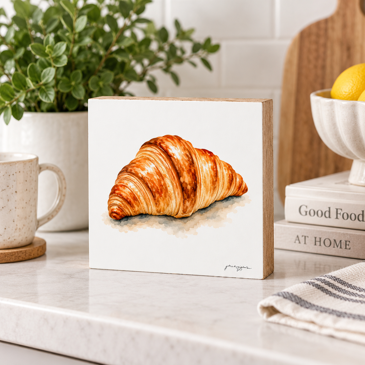 A square ceramic tile or coaster featuring a detailed watercolor illustration of a golden-brown croissant, displayed on a white marble kitchen countertop next to a mug, a potted plant, and books.