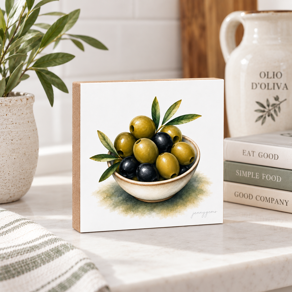 A square decorative art print or tile featuring a painted still life of green and black olives in a bowl, styled on a marble countertop with a potted plant, books, and a ceramic pitcher in a kitchen setting.