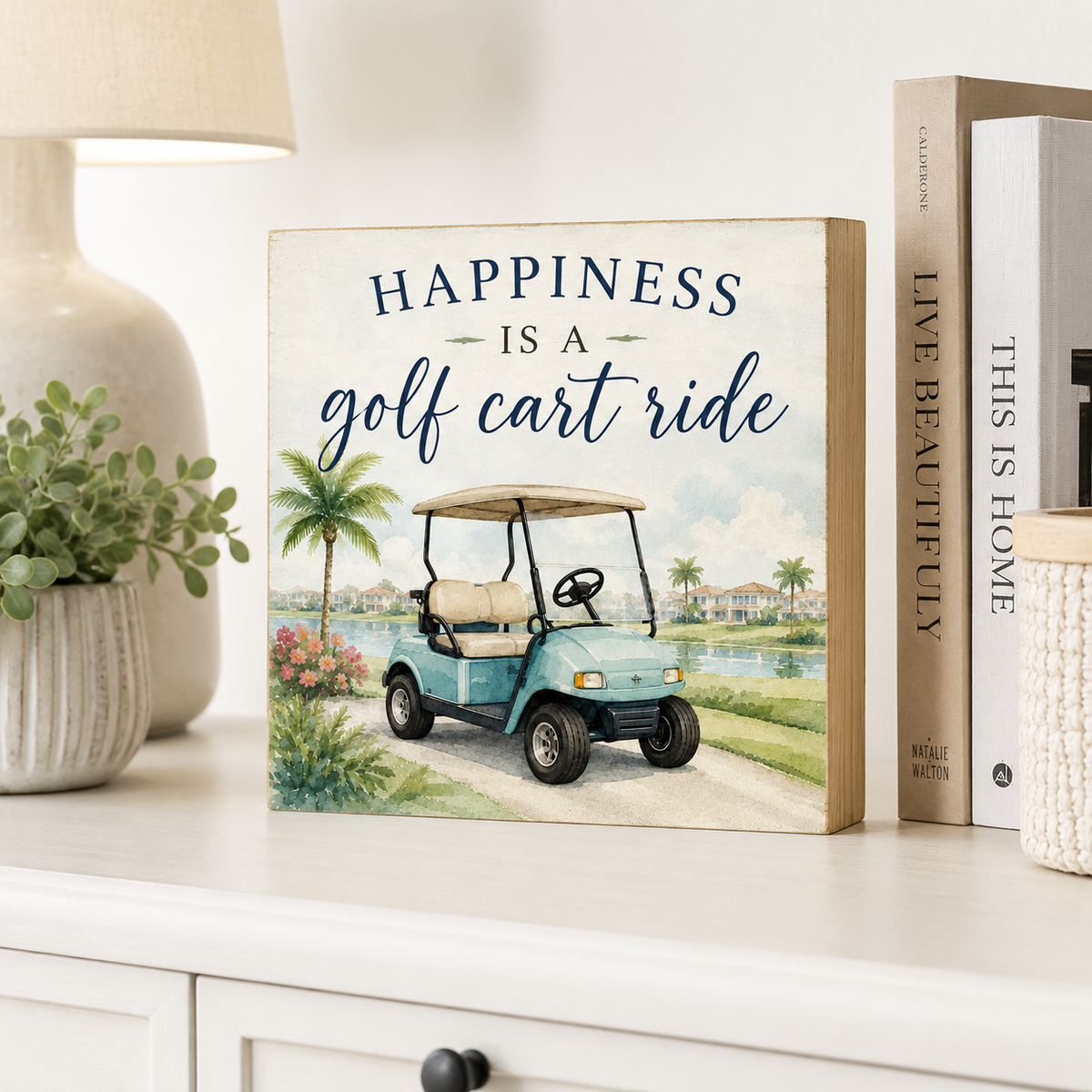 A square decorative sign with a golf cart illustration and the text 'HAPPINESS IS A golf cart ride' sits on a white cabinet beside a lamp, plant, and books.