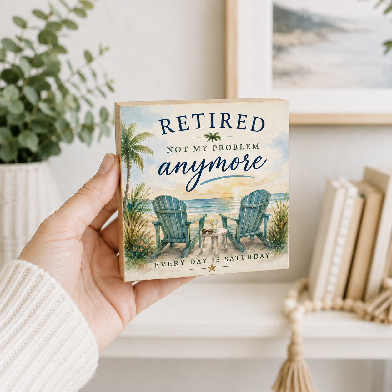 A hand holds a square decorative wooden sign featuring a beach scene with two Adirondack chairs and the text 'RETIRED NOT MY PROBLEM anymore' and 'EVERY DAY IS SATURDAY'. The background shows a cozy indoor setting with a plant, framed art, and books.