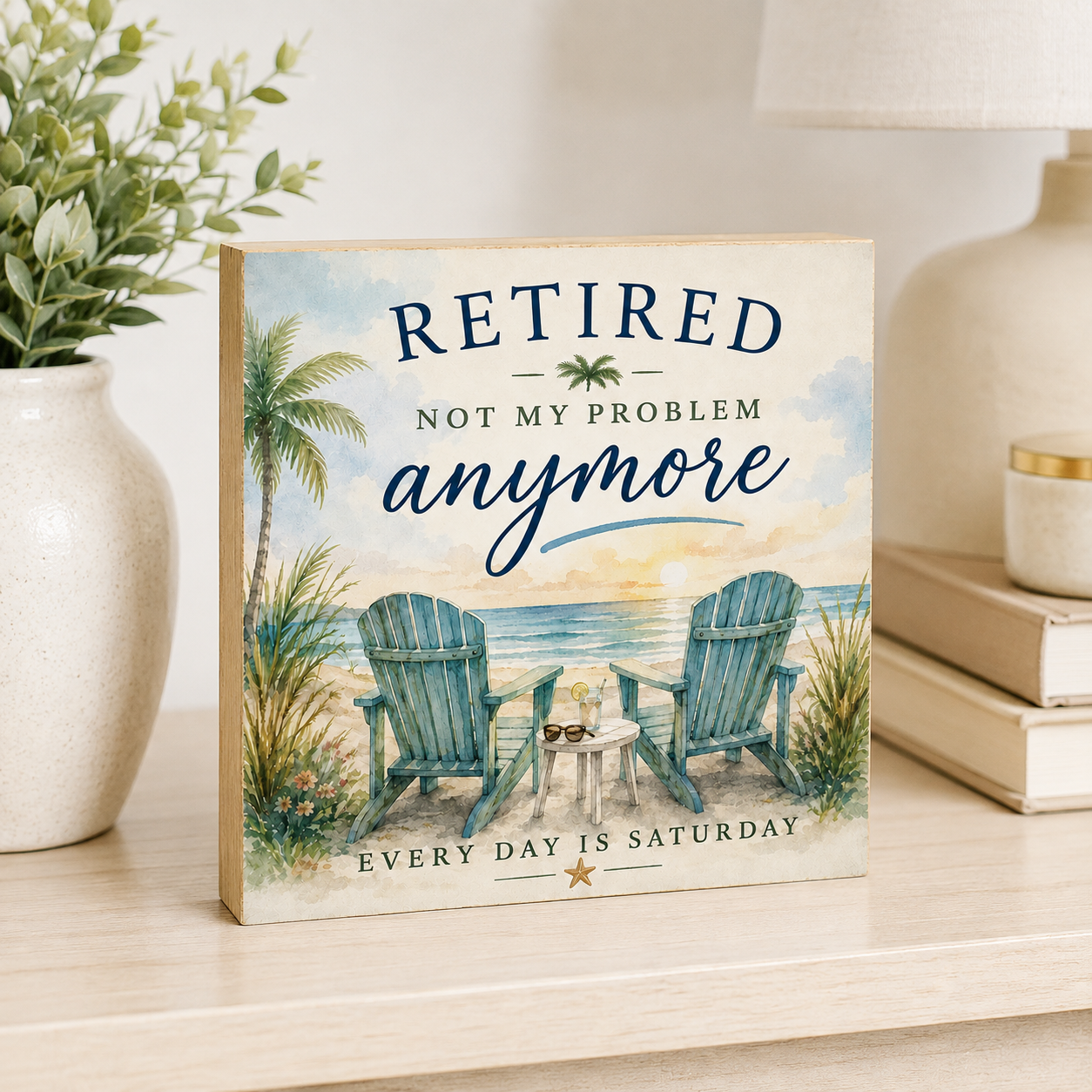 A square decorative sign with a beach scene illustration and the text 'RETired NOT MY PROBLEM anymore' and 'EVERY DAY IS SATURDAY' sits on a light wood shelf beside a vase with greenery and stacked books.