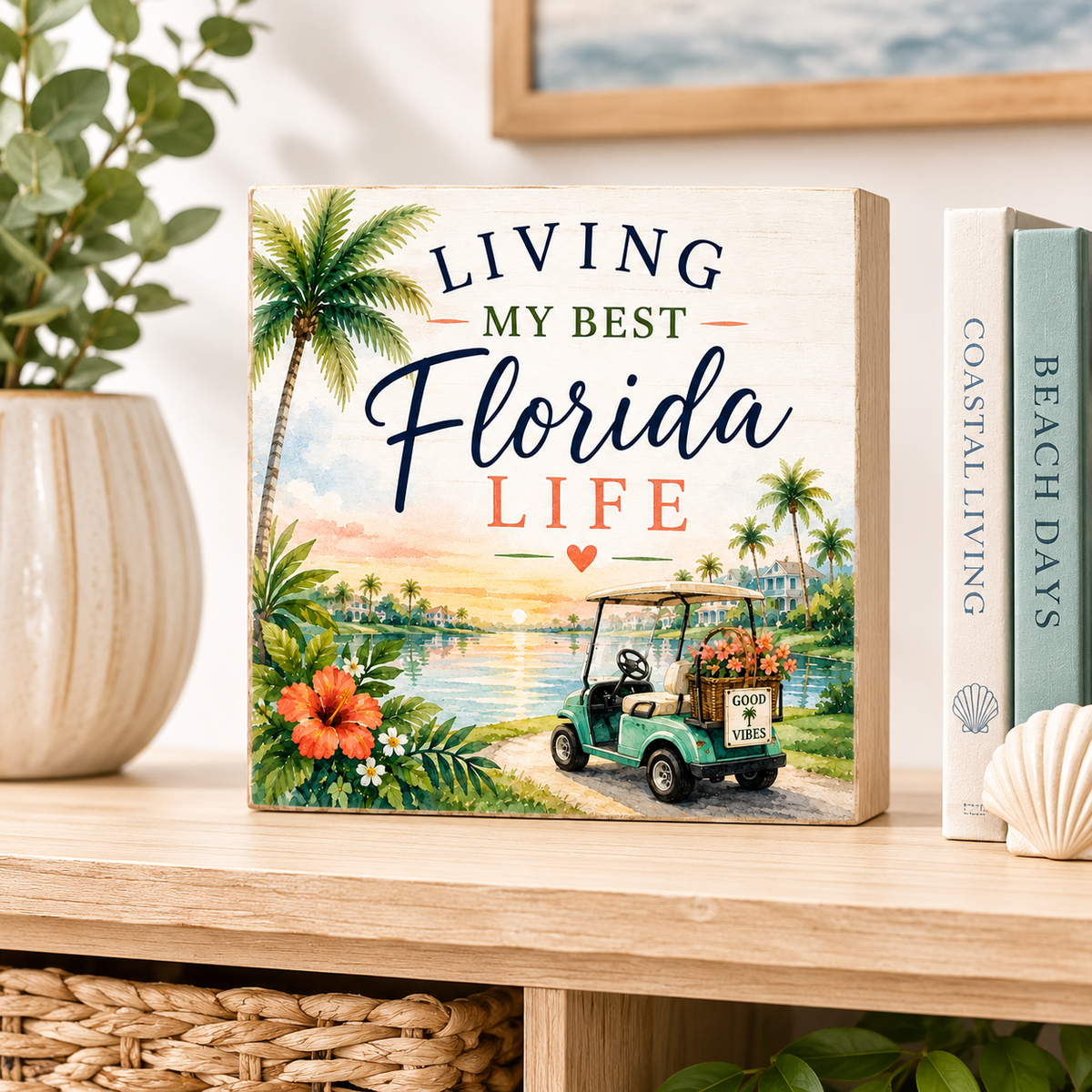A square decorative wooden sign with a tropical Florida scene and the text 'LIVING MY BEST Florida LIFE' sits on a light wood shelf beside books and a plant. The sign shows a palm tree, water, flowers, and a golf cart.