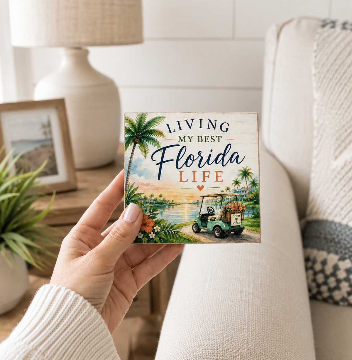 A hand holds a square decorative sign that reads 'Living My Best Florida Life' with a tropical beach scene, palm trees, water, and a golf cart. The background shows a cozy living room setting with a sofa, lamp, and framed picture.