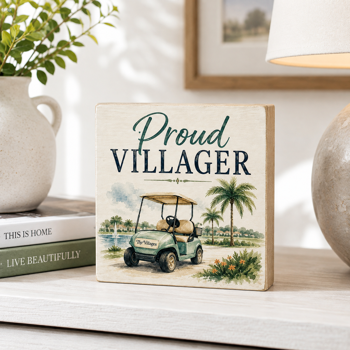 A square decorative wooden sign with the words 'Proud VILLAGER' and an illustration of a golf cart on a beach, styled on a tabletop next to books, a plant, and a lamp.