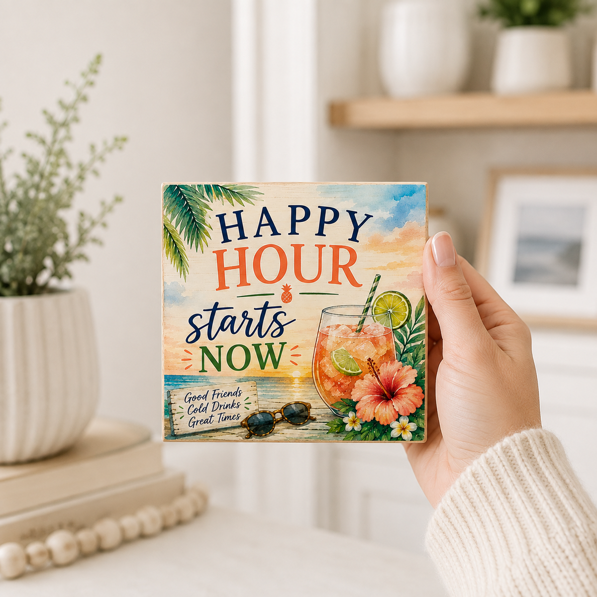 A hand in a white knit sleeve holds a square decorative coaster featuring a tropical beach scene and the text 'Happy Hour starts now' with a cocktail illustration. The background shows a softly blurred indoor setting with shelves, plants, and a framed picture.