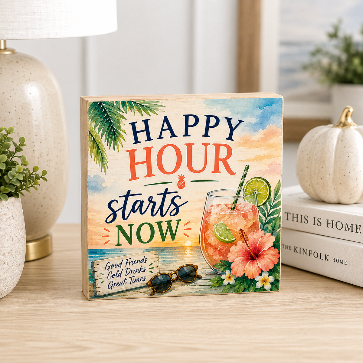 A square decorative wooden sign with a tropical beach scene and the text 'HAPPY HOUR starts NOW' sits on a light wood table. The sign features a colorful drink, hibiscus flower, sunglasses, palm leaves, and a small boat.