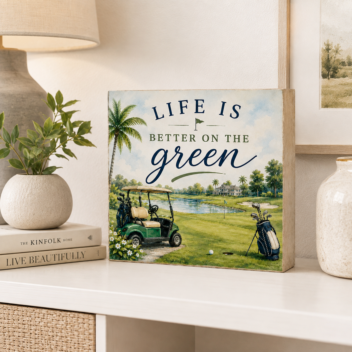 A square decorative wooden sign with a golf course scene and the text 'LIFE IS BETTER ON THE green' displayed on a white shelf beside a lamp, books, and vases.