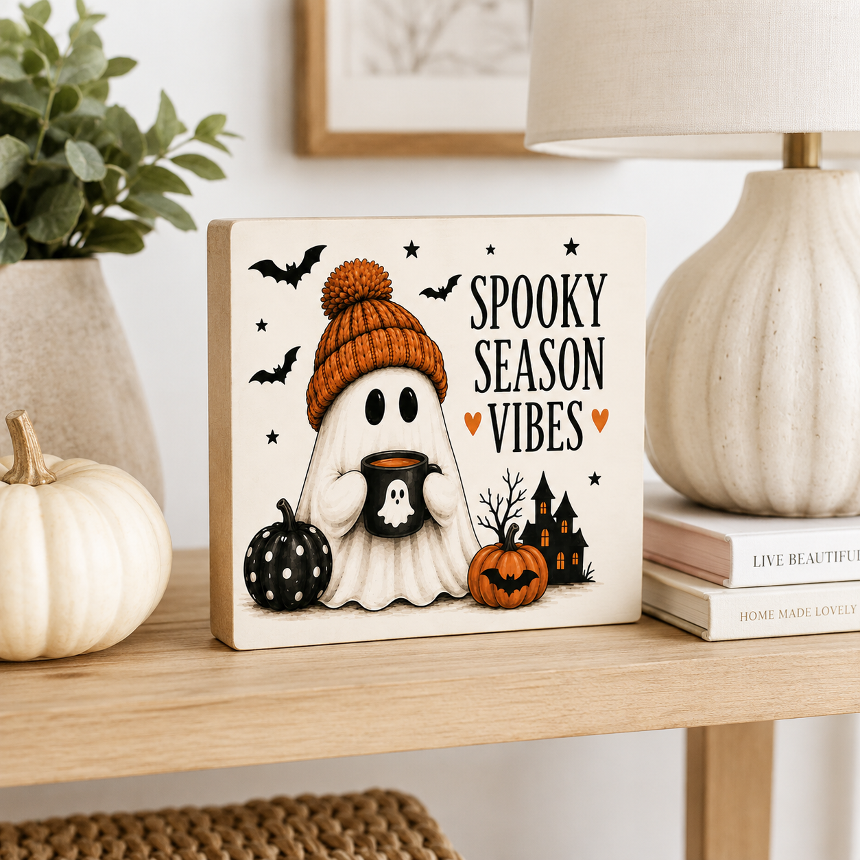 A square decorative wooden sign with a Halloween illustration reading 'SPOOKY SEASON VIBES,' styled on a wooden shelf beside a white pumpkin, books, a plant, and a lamp.