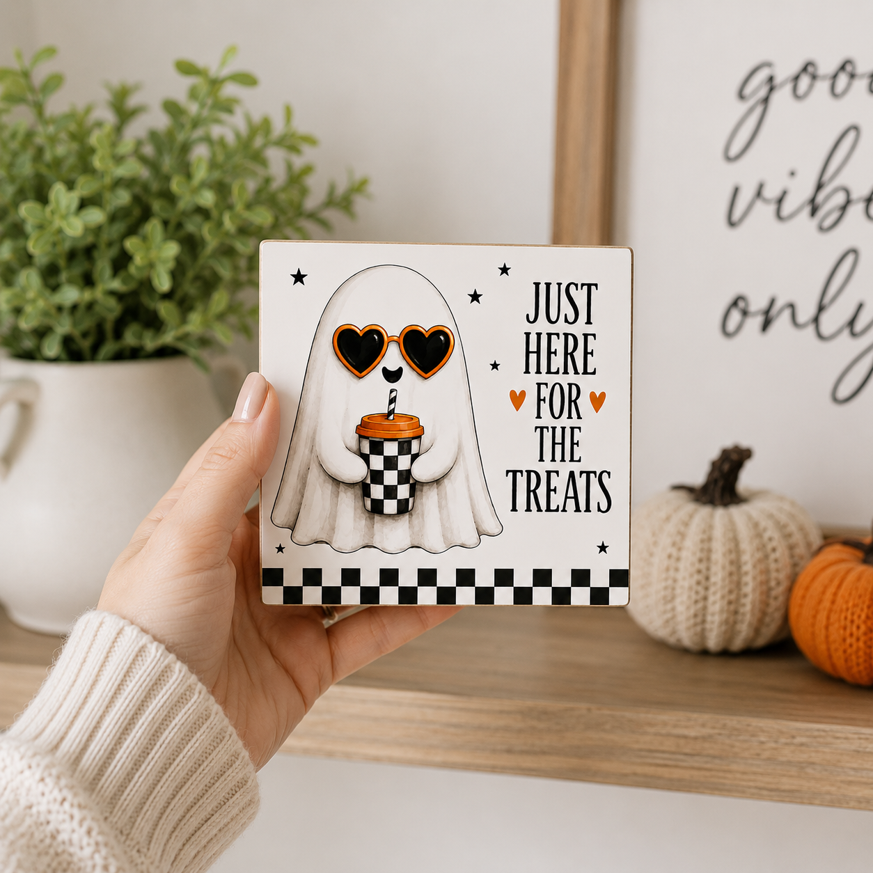 A hand holds a square white plaque featuring a cartoon ghost wearing heart-shaped sunglasses and holding an orange pumpkin cupcake, with the text 'JUST HERE FOR THE TREATS' and a black-and-white checkered border. The background shows a cozy indoor setting with a potted plant, a knitted pumpkin, and a framed sign.
