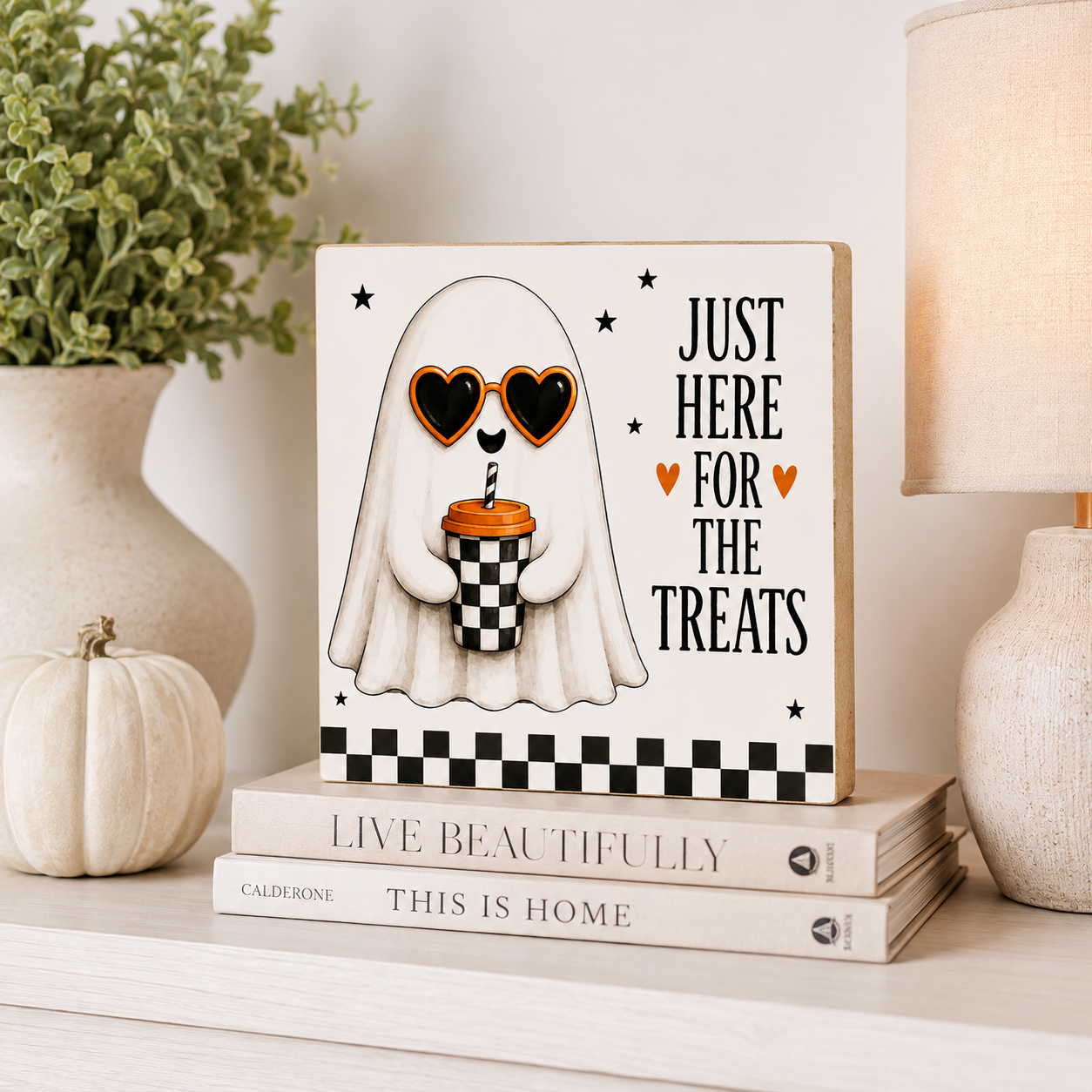 A square decorative sign featuring a cartoon ghost with heart-shaped sunglasses holding a drink, next to the text 'JUST HERE FOR THE TREATS' with small orange hearts. The sign is styled on a shelf with books, a small pumpkin, a plant, and a lamp.