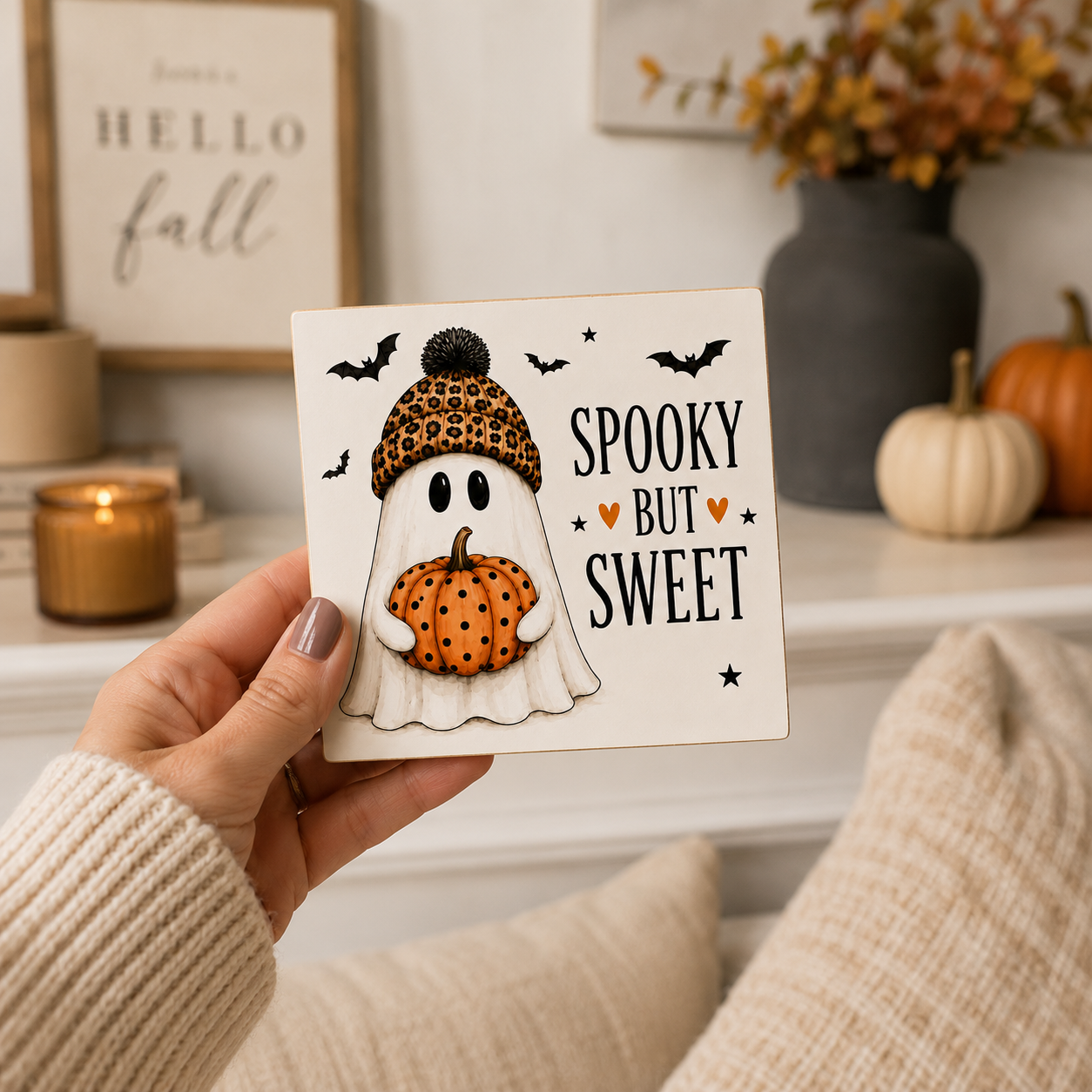 A hand holds a square decorative sign that reads 'Spooky But Sweet' with a ghost character wearing a leopard-print beanie and holding an orange pumpkin. The scene is a cozy indoor setting with fall decor, including candles, framed wall art, and pumpkins.