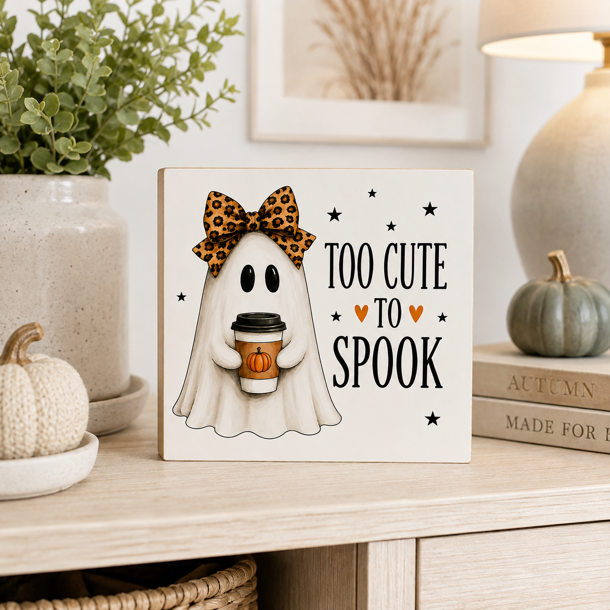 A square decorative sign with a cartoon ghost holding a pumpkin coffee cup and wearing a leopard-print bow, next to the text 'TOO CUTE TO SPOOK' with small hearts and stars. The sign is styled on a light wood shelf with a potted plant, books, and a small pumpkin.