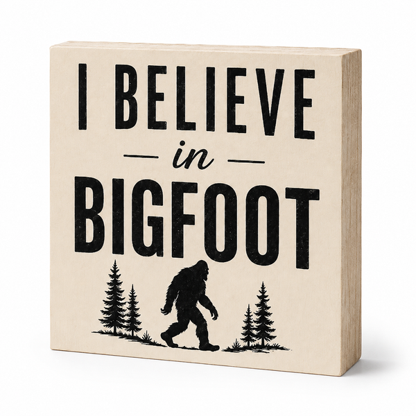 A square decorative block sign with a light beige wood-grain finish and bold black text reading 'I BELIEVE in BIGFOOT,' plus a black silhouette of a Bigfoot figure between pine trees, shown on a clean white background.